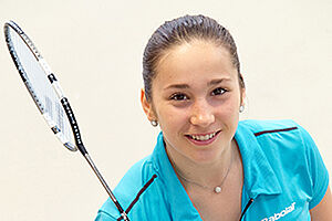 Jeune femme brune souriante, vêtue d'un tee-shirt bleue et arborant une raquette de badminton.