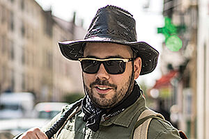 Homme brun, barbu, souriant, portant un chapeau, des lunettes de soleil, une chemise et à sac à dàs d'aventurier