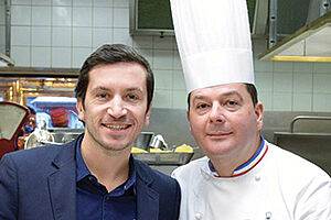 Jeune hommes brun posant au côté d'un cuisinier portant sa toque, dans la cuisine du restaurant de Paul Bocuse.