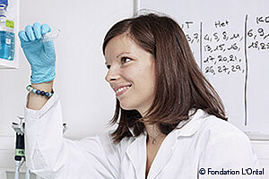 Jeune femme brune, assise, portant une blouse blanche et des gants bleus observant de petites éprouvettes