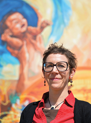 Une jeune femme brune aux cheveux courts, portant des lunettes et posant devant une fresque colorée.