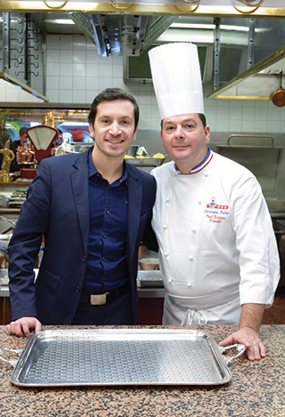 Jeune homme brun posant au côté d'un cuisinier portant sa toque, dans la cuisine du restaurant de Paul Bocuse.