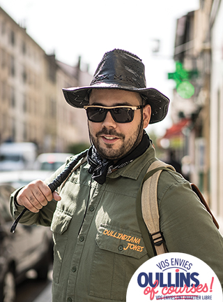 Homme brun, barbu, souriant, portant un chapeau, des lunettes de soleil, une chemise et à sac à dàs d'aventurier